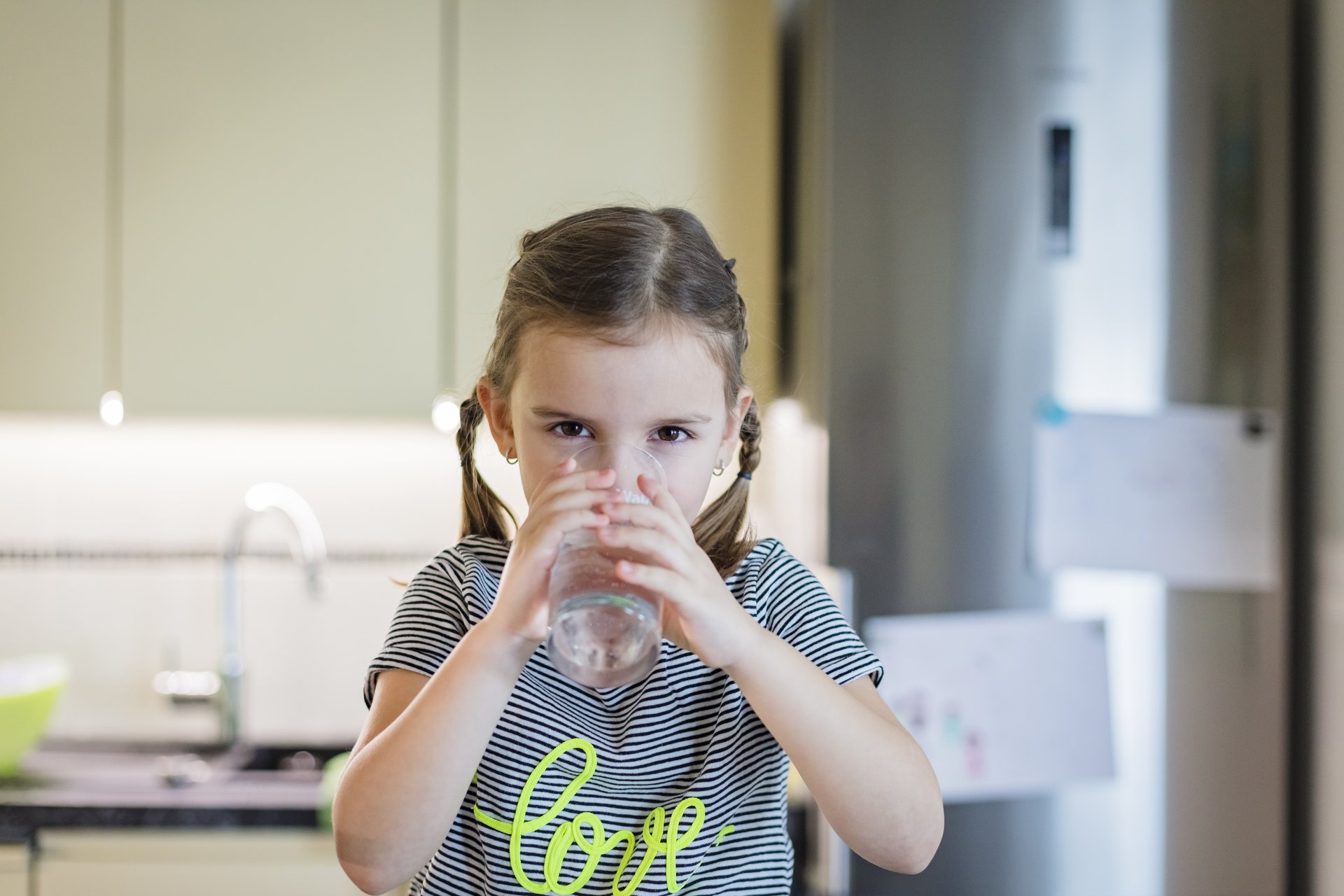 Ein kleines Mädchen mit Zöpfen trinkt natürliches Mineralwasser von Waldquelle in der Küche.