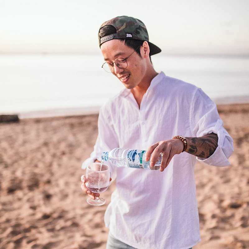 Ein junger Mann am Strand füllt sein Glas mit natürlichem ALPQUELL Mineralwasser.