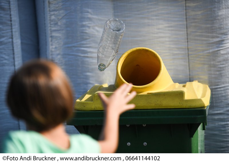 Ein Kind wirft eine PET-Flasche zum Recyceln in einen gelb-grünen Sammelcontainer. 