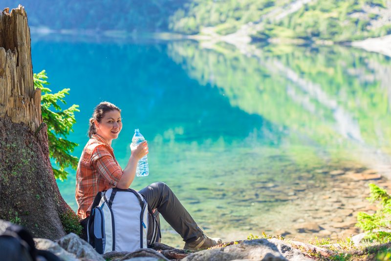 Am Rande eines klaren Sees sitzt eine Frau mit einer Flasche natürlichem Mineralwasser.